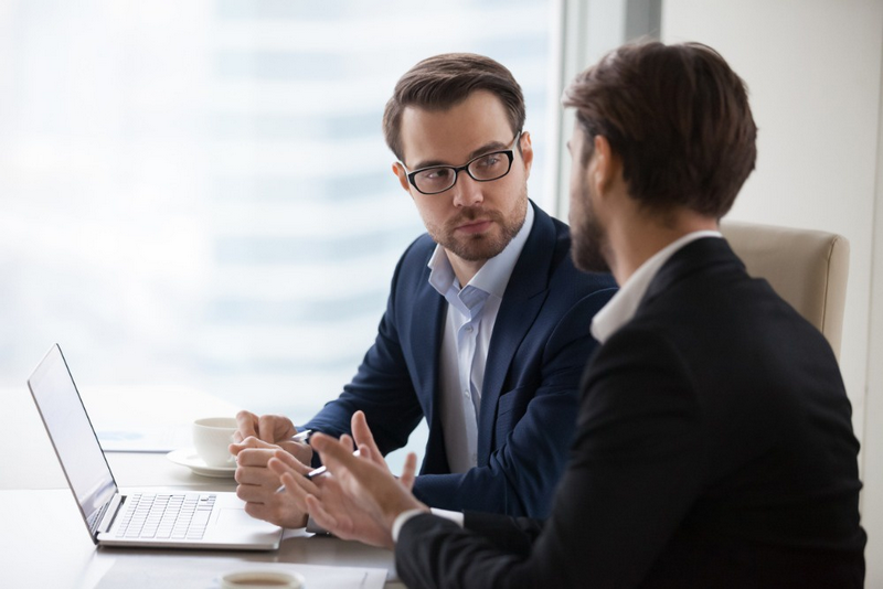 Two business professionals having a discussion at a table with a laptop open between them.