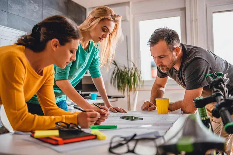 Three colleagues collaborating on a project, reviewing plans on a table in a bright office.