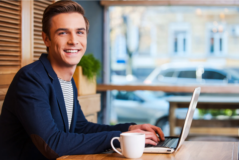 A young man with a cheerful expression working on a laptop at a café table, with a coffee cup beside him.